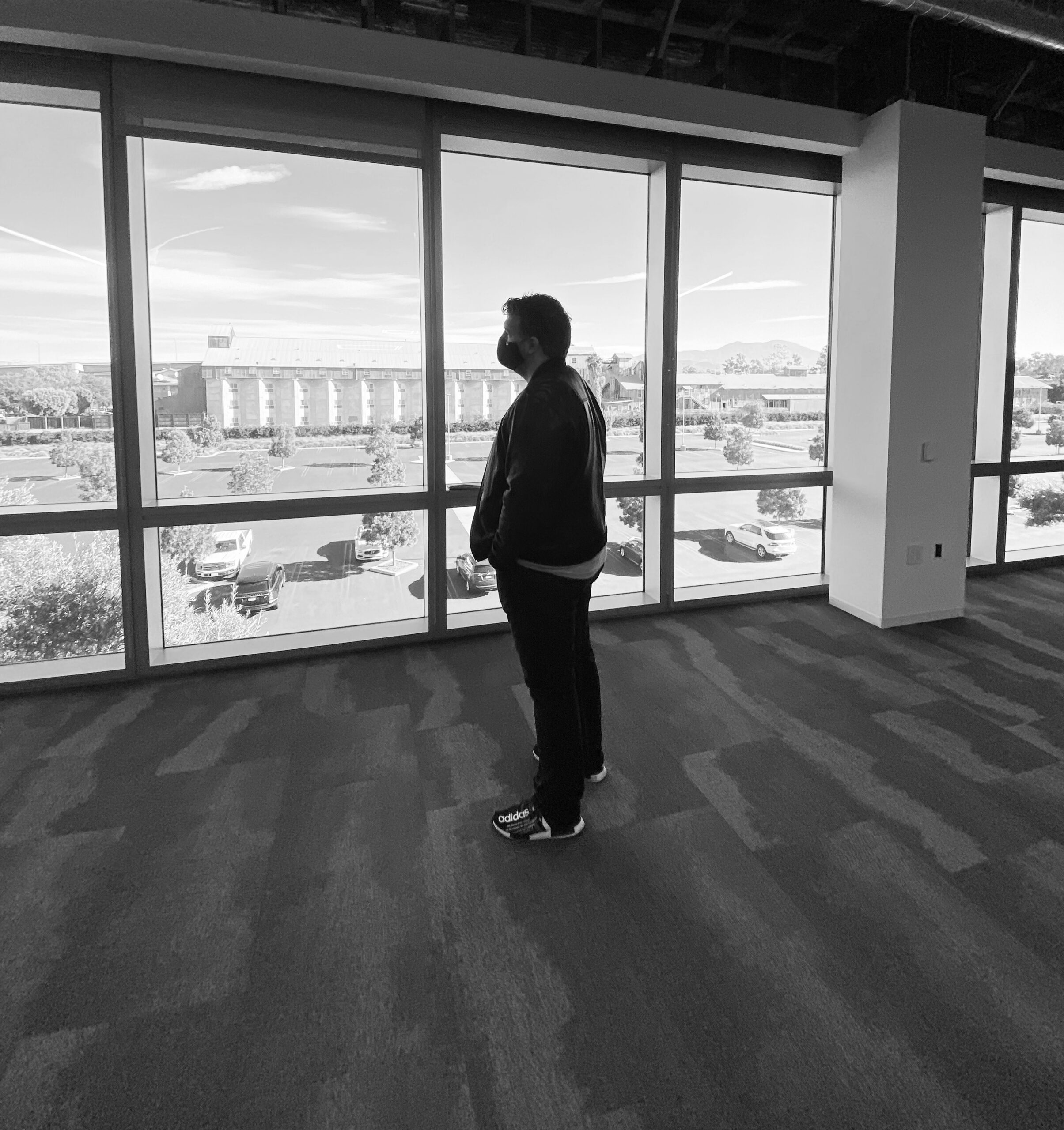 A man stands alone in an empty, unfinished office, hands in his pockets, looking out floor-to-ceiling windows over an empty parking lot.