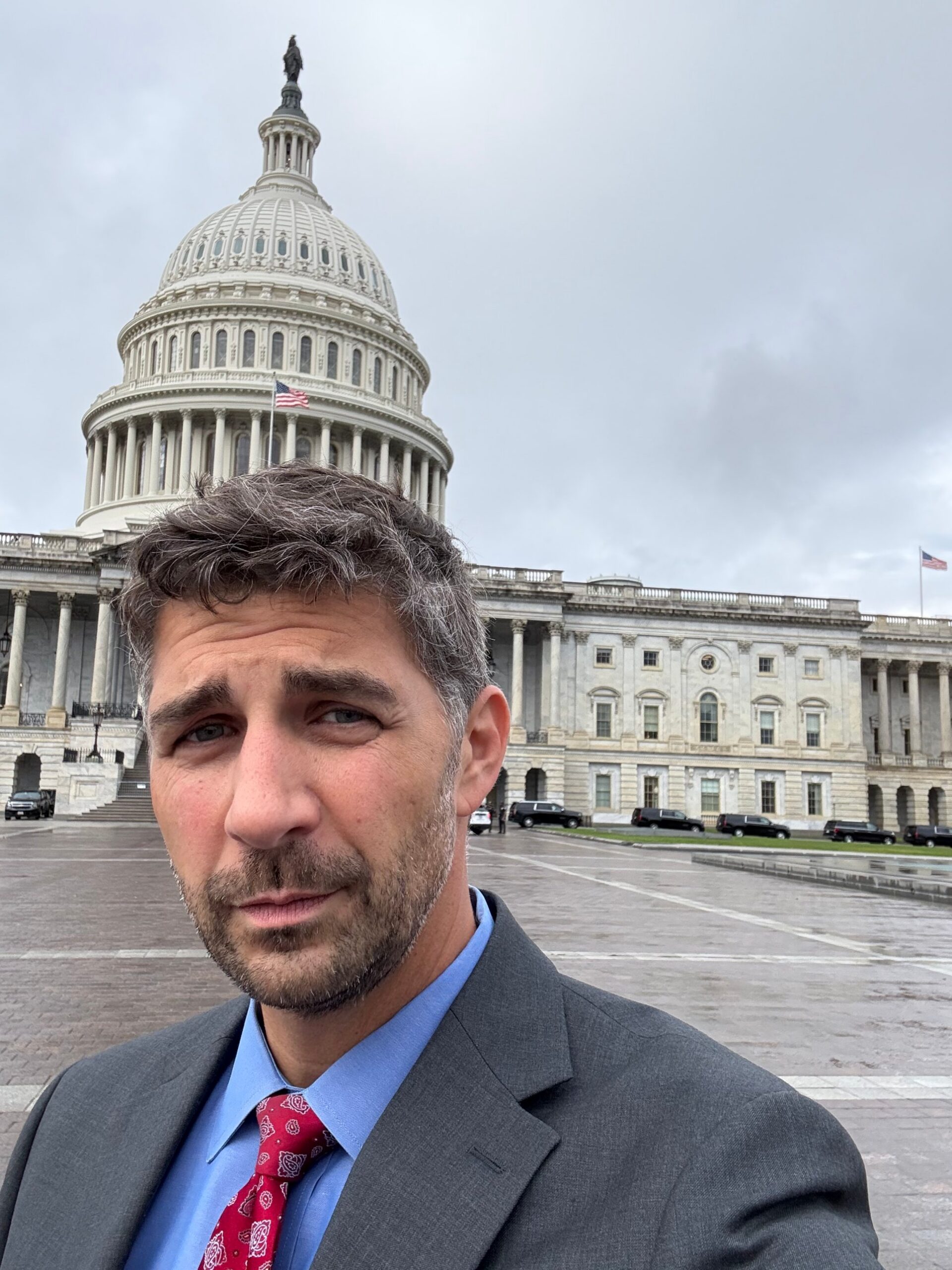 Pierre Rogers in a suit and tie standing in front of the United States Capitol building on an overcast day.